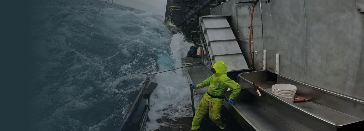 Fisherman in bright yellow rain gear working on a fishing boat deck amidst rough ocean waves during a stormy sea.